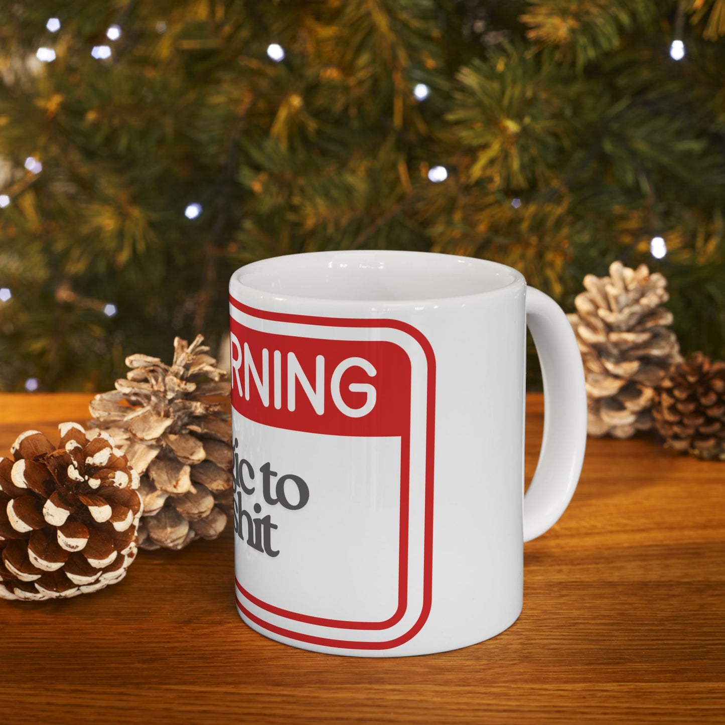 White mug with a red and white warning label design, placed on a wooden surface with pine cones and a Christmas tree in the background.